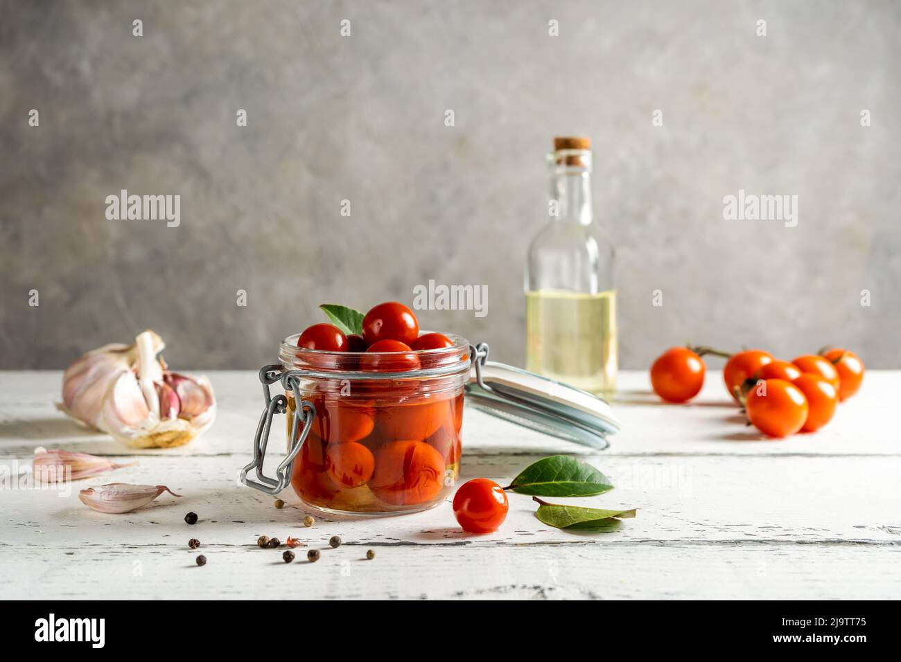 Eingelegte Tomaten in einem Glas mit Knoblauch, Öl, frischen Tomaten, Pfeffer und Lorbeerblättern auf weißem Holztisch mit grauem Hintergrund Stockfoto