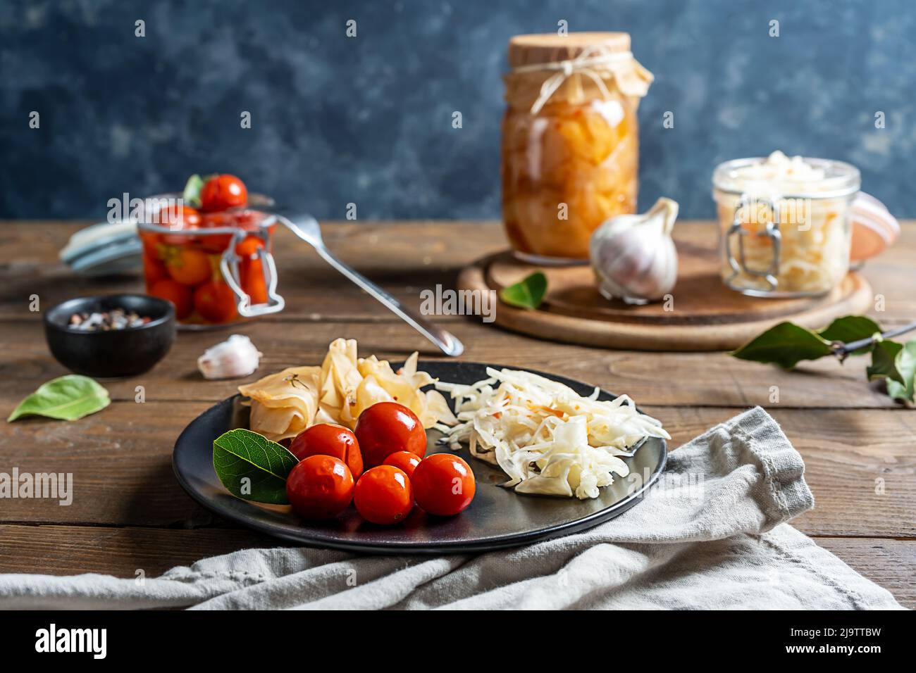 Eingelegte Tomaten, Kimchi, Kohl im Glas auf Schneidebrett und Teller mit Serviette und Gabel auf Holztisch mit grauem Hintergrund Stockfoto