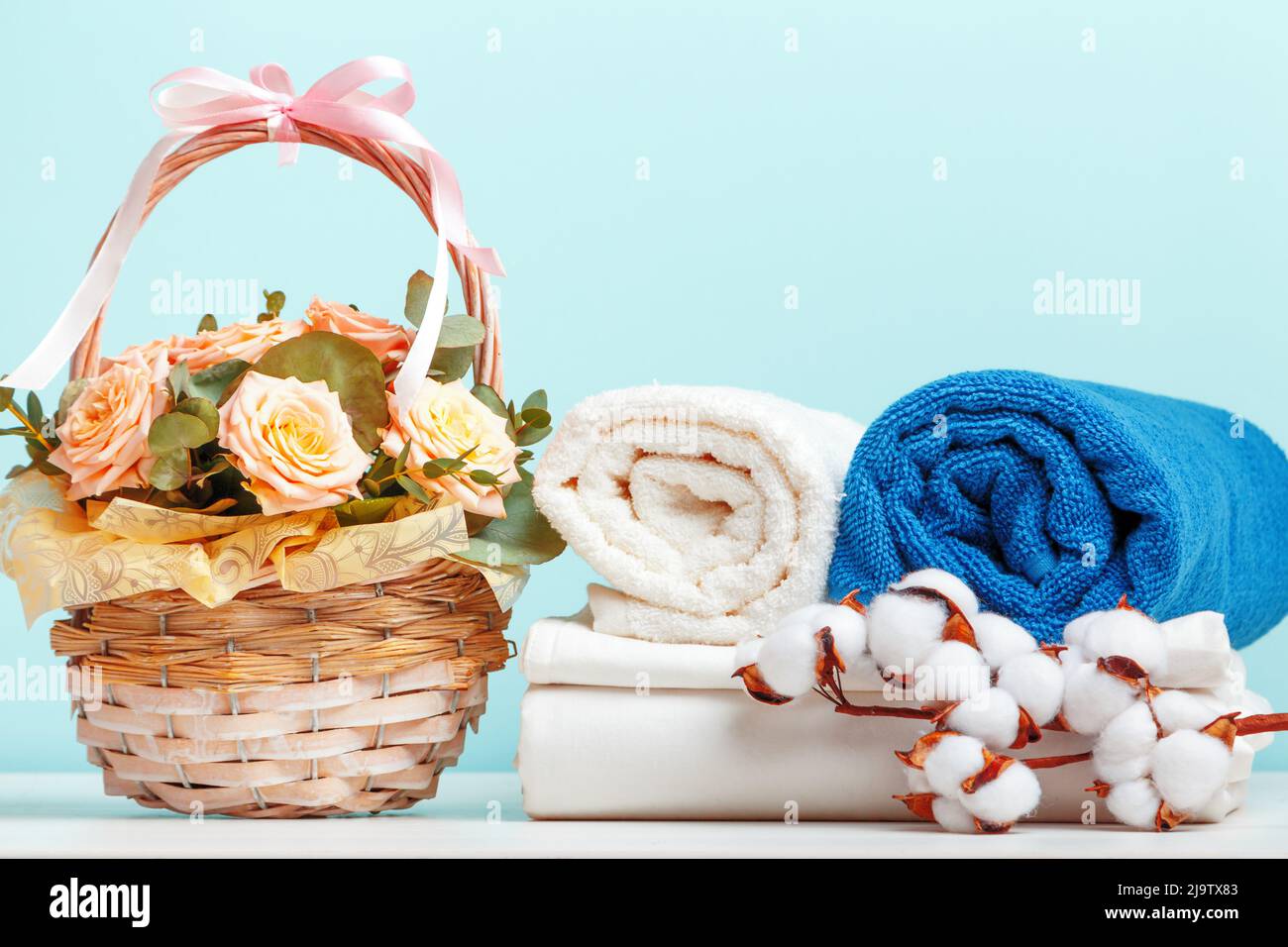 Ein Stapel weißer Bettwäsche, Bettwäsche und eine Rolle Handtücher. Ein Korb mit Blumen und ein Zweig aus Baumwolle auf dem Tisch. Stockfoto
