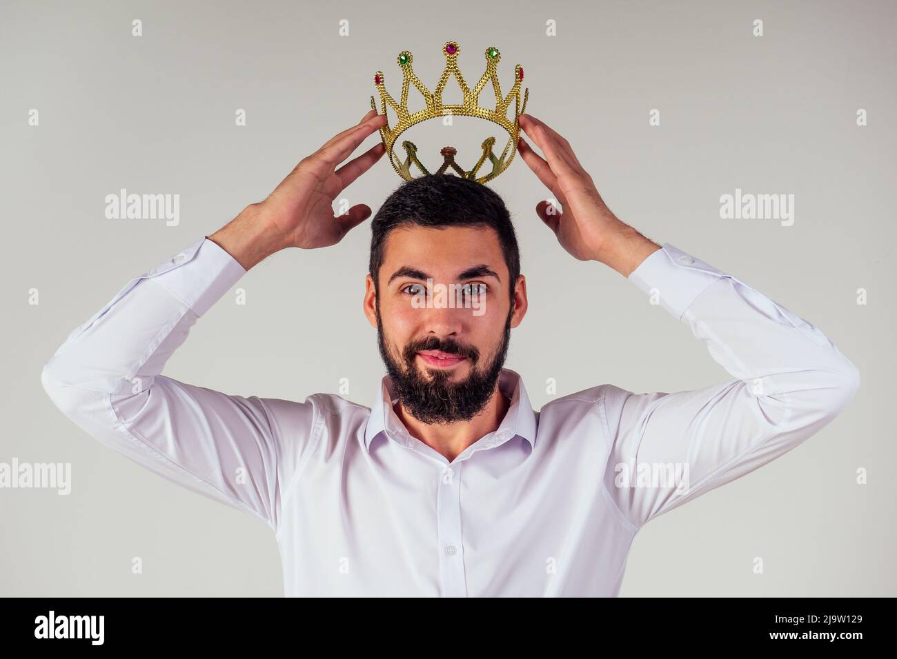 Porträt eines lächelnden bärtigen Geschäftsmanns mit einer goldenen Krone auf dem Kopf auf weißem Hintergrund, aufgenommen im Studio Stockfoto