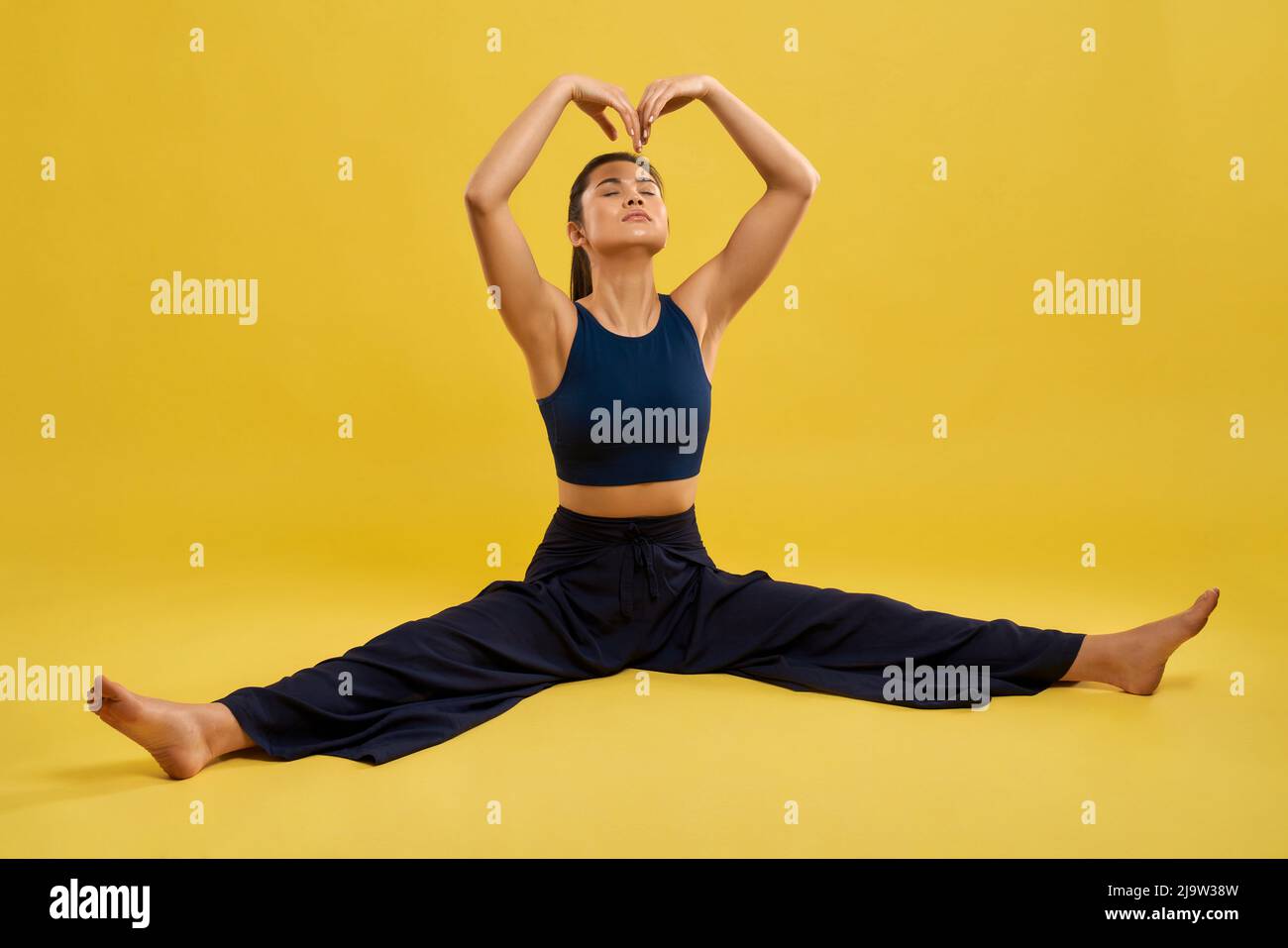 Weibliche Yoga-Coach tut Split, Dehnung der Muskeln während der Übung in der Halle. Vorderansicht eines schlanken Mädchens, das im Garn sitzt und während des Yoga-Trainings die Arme über den Kopf hält, isoliert auf Gelb. Konzept des Yoga. Stockfoto