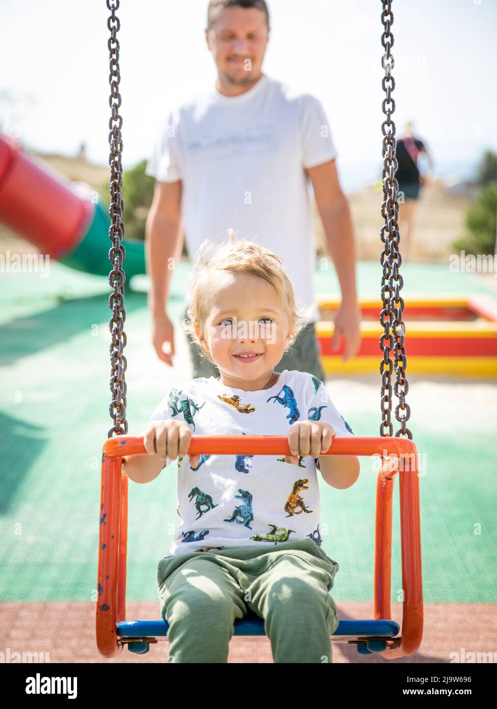 Ein kleiner kaukasischer blonder Junge, 3 Jahre alt, schwingt an einem sonnigen, warmen Sommertag auf dem Spielplatz von seinem jungen Vater auf einer Schaukel. Stockfoto