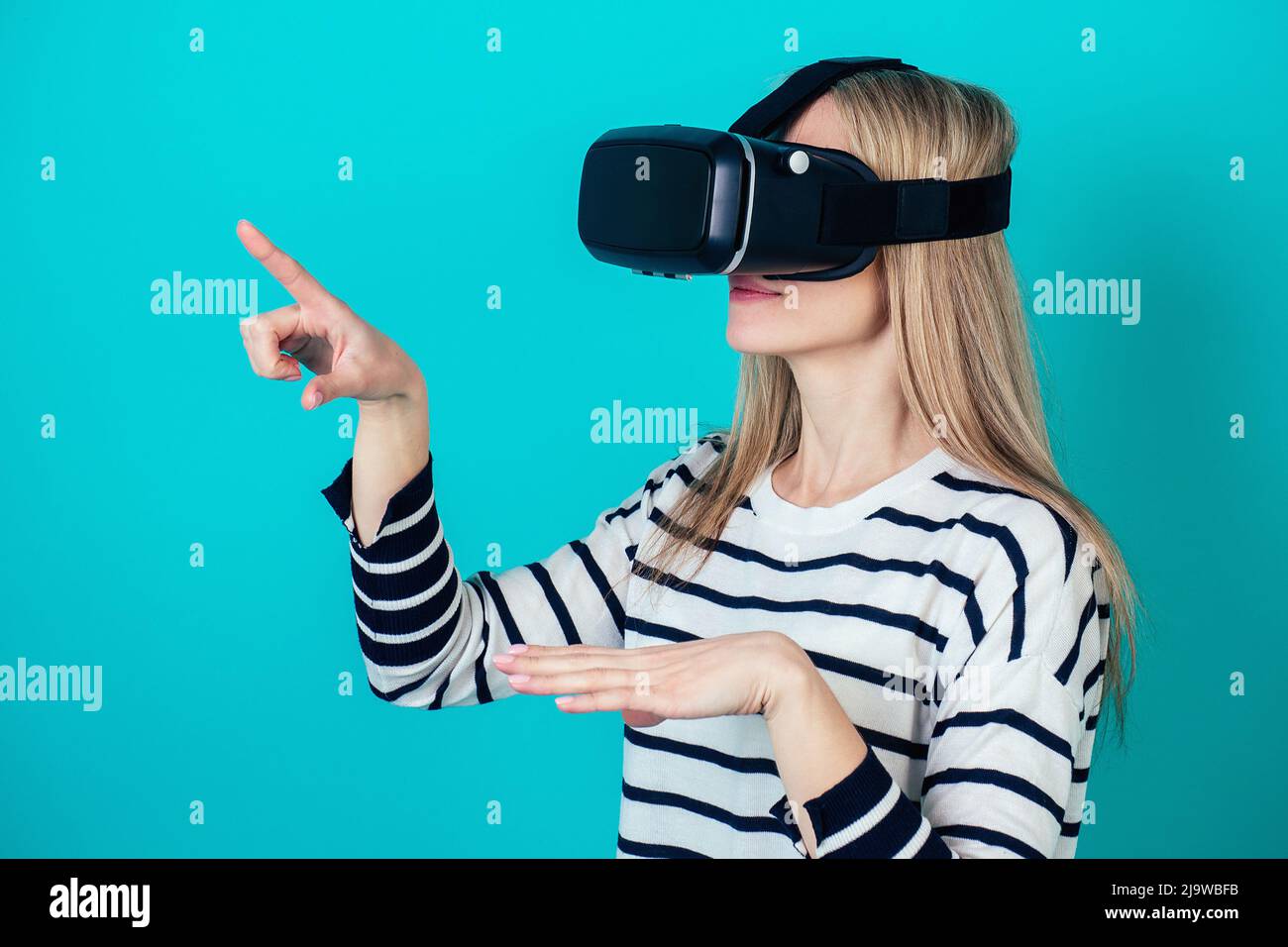 Attraktive Frau in einem Helm (Brille) der virtuellen Realität berührt Hände zeigt Finger auf einem blauen Hintergrund im Studio. Das Konzept der Zukunft Stockfoto