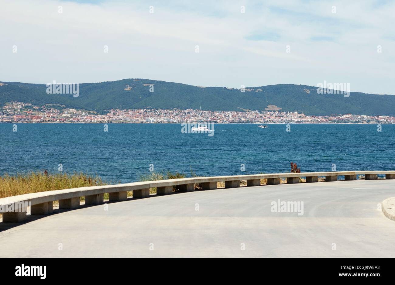 Vorbei an einer asphaltierten Straße entlang der Küste. Ein Boot fährt in der Ferne vor der Kulisse eines Küstenortes. Konzentrieren Sie sich auf das Boot Stockfoto