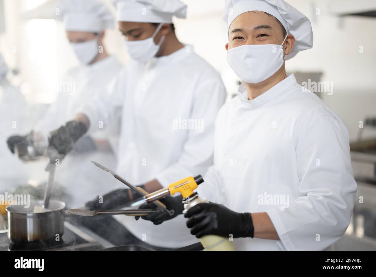 Multirassisches Team von Köchen kocht in weißer Uniform zusammen in der Küche. Asiatischer Koch, der Fleisch mit Brenner, lateinamerikanische und europäische Küche auf dem Hintergrund braten lässt. Köche tragen Gesichtsmasken und Schutzhandschuhe Stockfoto