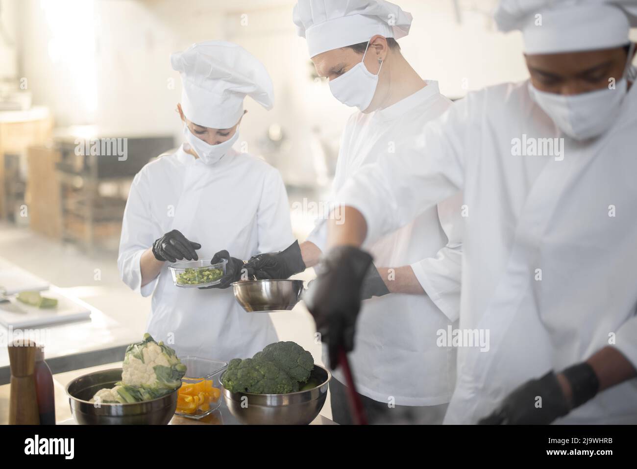 Multirassisches Team von Köchen kocht in weißer Uniform zusammen in der Küche. Asiatische, lateinische und europäische Jungs kochen zusammen. Köche tragen Gesichtsmasken und Schutzhandschuhe Stockfoto