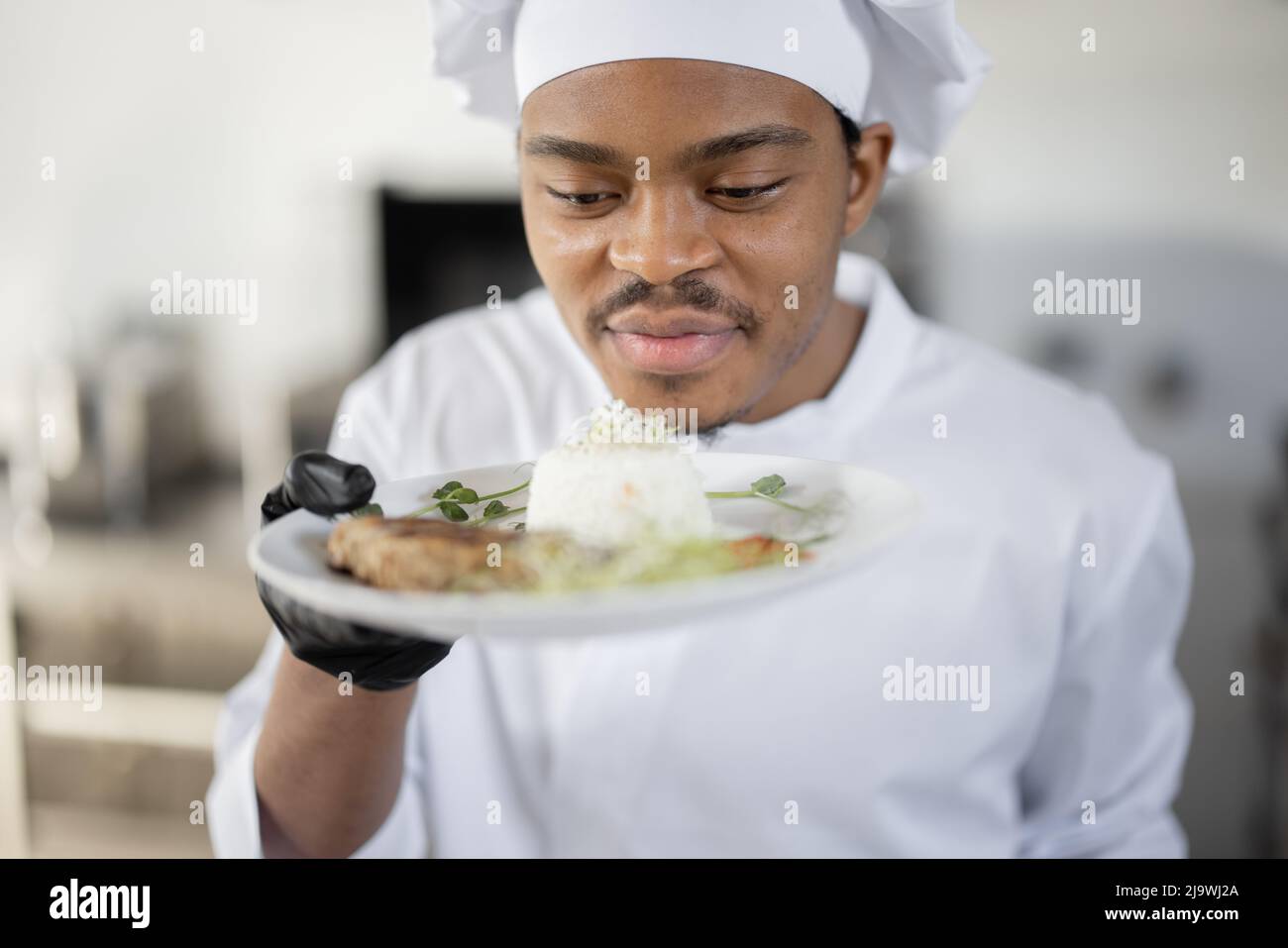 Porträt eines jungen, gutaussehenden, lächelnden Küchenchefs in Uniform, der mit einer Fertigmahlzeit in der Küche steht. Fröhlicher lateinamerikanischer Koch, der im Restaurant arbeitet. Konzept der beruflichen Tätigkeit und Haute Cuisine Stockfoto