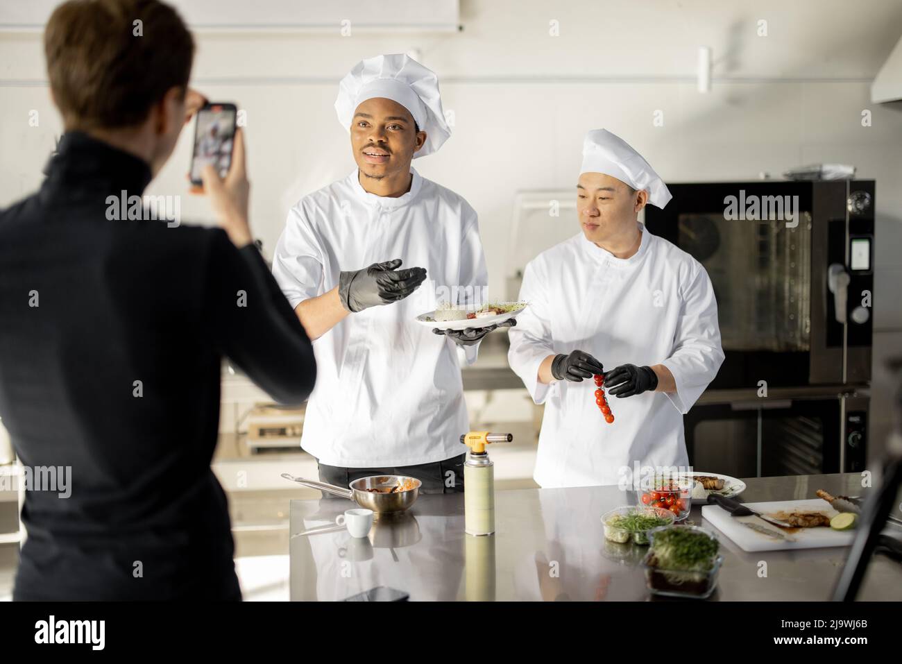 Mann zeichnet am Telefon den Prozess des Kochens von zwei Köchen in einer professionellen Küche auf. Lateinamerikanische und asiatische Köche bereiten Speisen für soziale Medien zu. Konzept des kulinarischen Videobloggens Stockfoto