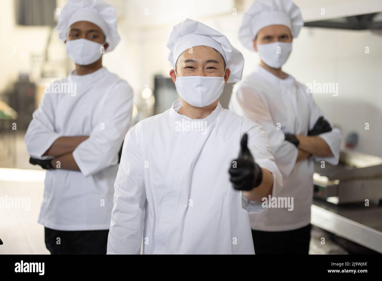 Porträt von drei gut gekleideten Köchen mit unterschiedlichen Ethnien, die in der Restaurantküche zusammenstehen. Asiatischer Koch mit OK-Zeichen, lateinische und europäische Männer im Hintergrund. Köche mit Gesichtsmasken, bereit, während einer Pandemie zu kochen Stockfoto