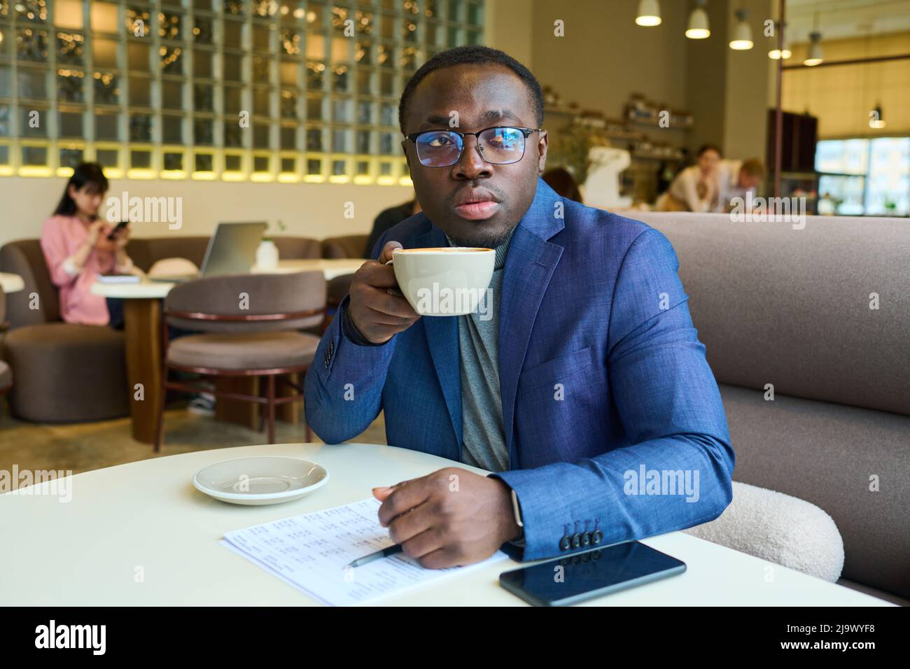 Afrikanischer junger Geschäftsmann im Anzug, der Kaffee trinkt, während er mit Dokumenten und Handy am Tisch im Café sitzt Stockfoto