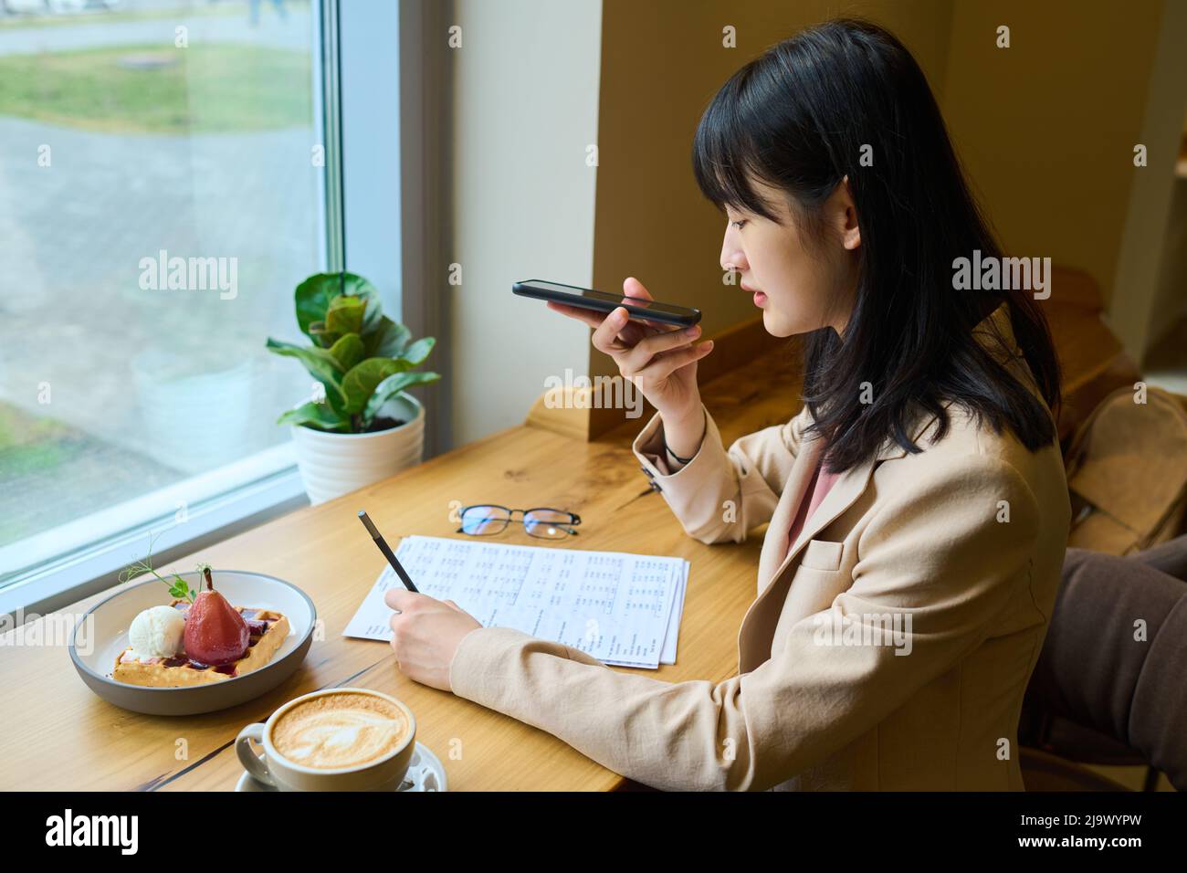 Junge Geschäftsfrau liest Dokumente und zeichnet Sprachnachrichten auf ihrem Mobiltelefon auf, während sie im Café frühstückt Stockfoto