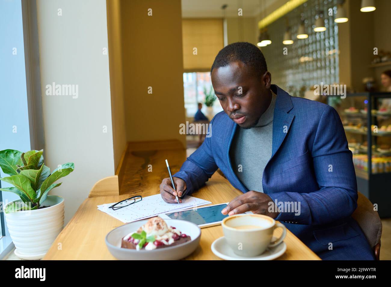 Afrikanischer Geschäftsmann, der sich auf IS konzentriert, arbeitet auf einem digitalen Tablet, berührt den Bildschirm und füllt Dokumente am Tisch mit Dessert im Café aus Stockfoto