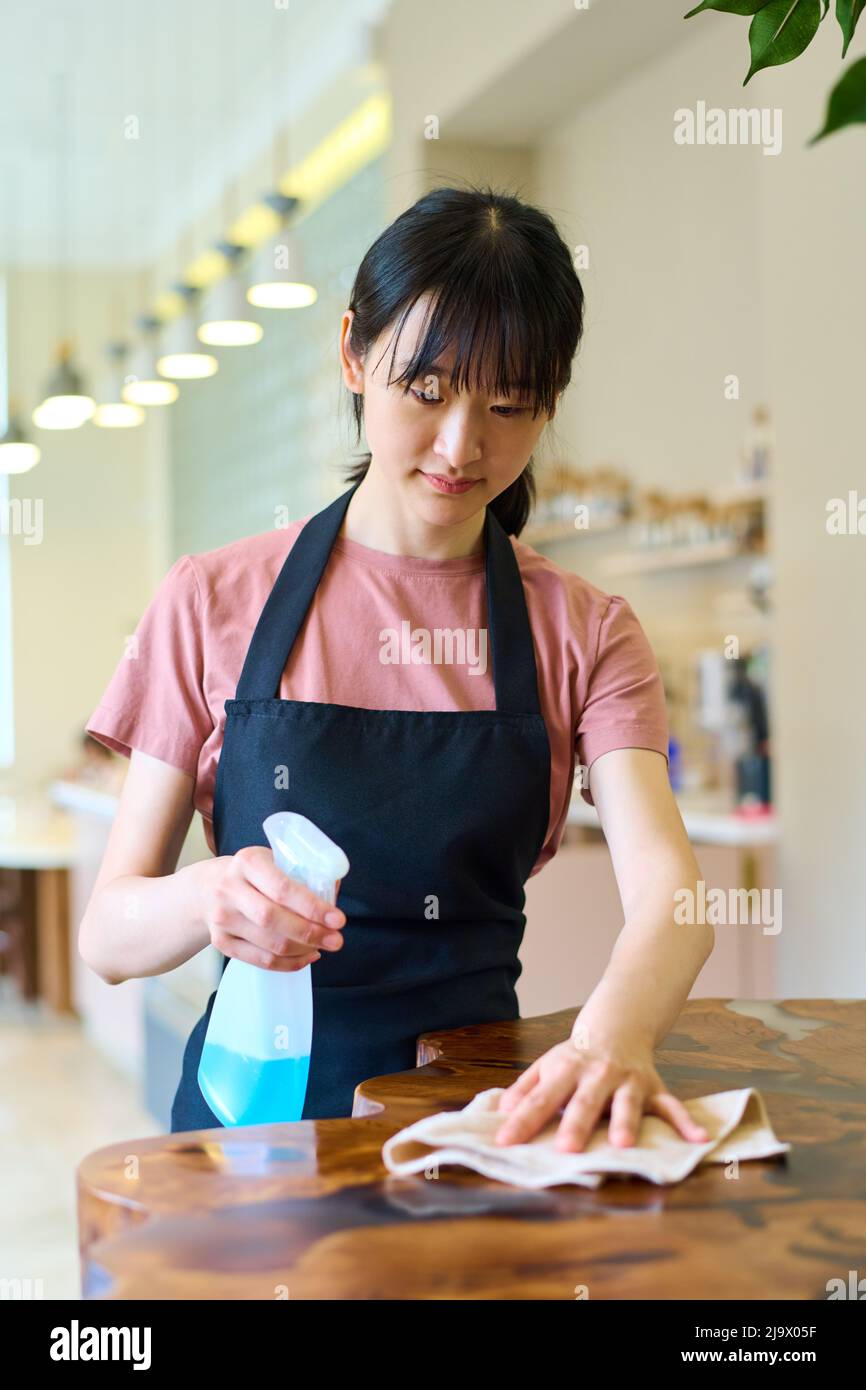 Junge asiatische Kellnerin in Schürze Wischtisch mit Reinigungsmittel vor dem Öffnen des Cafés Stockfoto