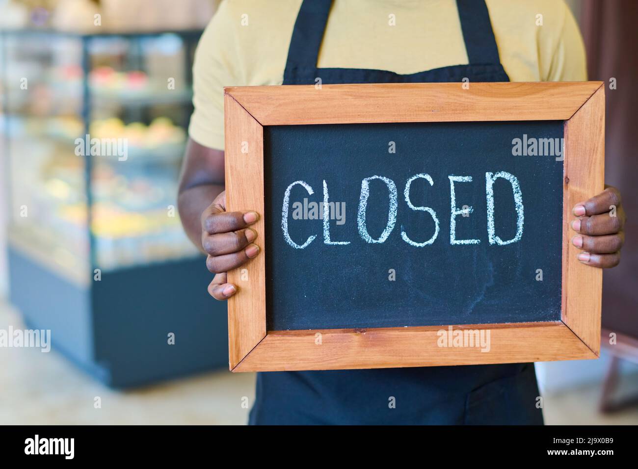 Nahaufnahme eines afroamerikanischen Kellners in Uniform, der das Brett mit geschlossenem Schild hält, während er im Café steht Stockfoto