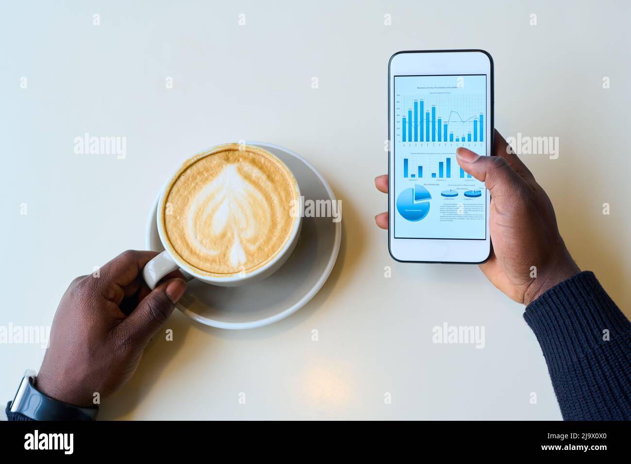 Blick aus der Perspektive eines afrikanischen Geschäftsmannes, der die Finanzmärkte auf dem Mobiltelefon untersucht, während er Cappuccino trinkt Stockfoto