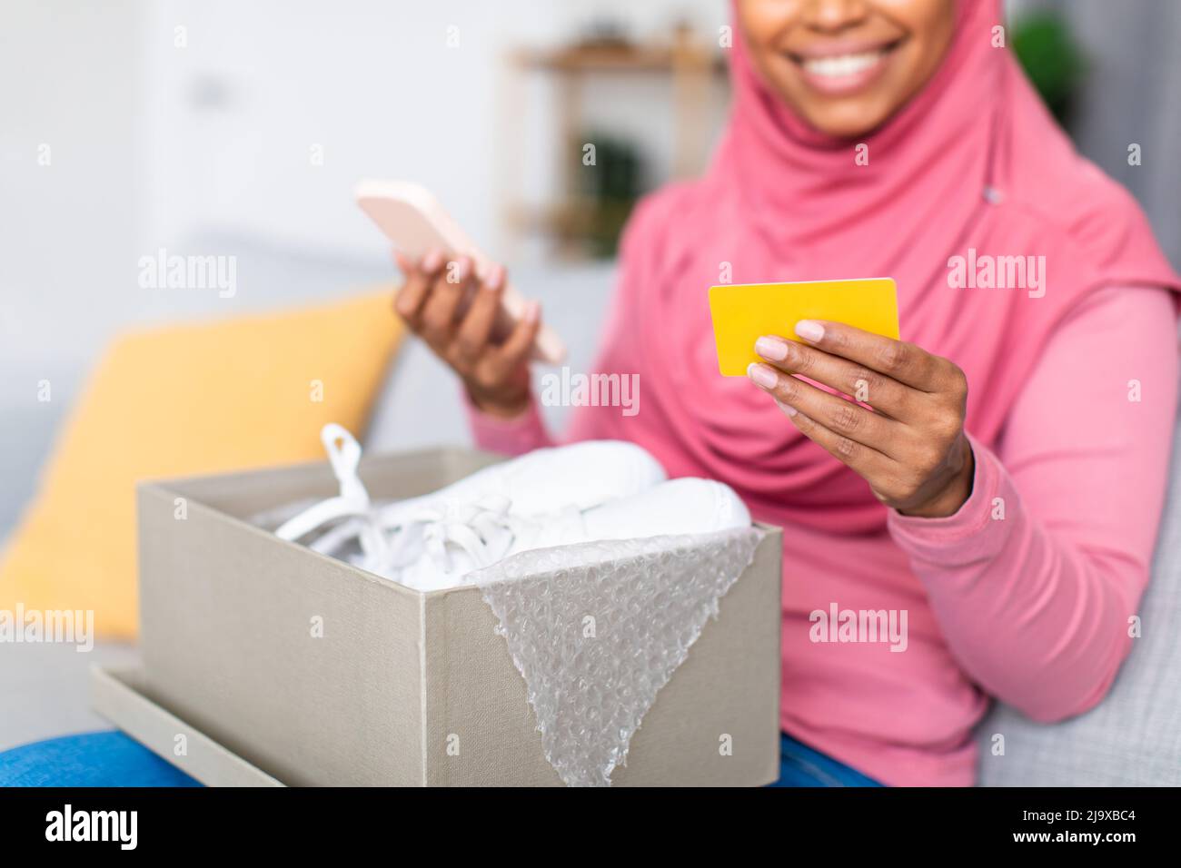 Aufgeregte afroamerikanische muslimische Dame mit Kreditkarte und Handy, die Karton mit neuen Schuhen auspackte Stockfoto