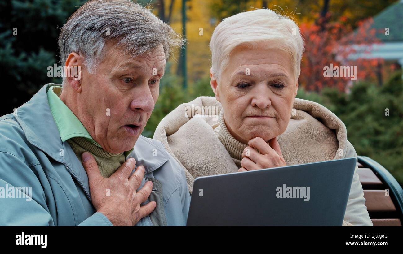 Schockiert alten Ehepaar Blick auf Laptop-Bildschirm sitzen im Herbst Park verärgert älteren Mann und Frau lesen schlechte Nachrichten besorgt Ehepartner im Alter fühlen Stockfoto