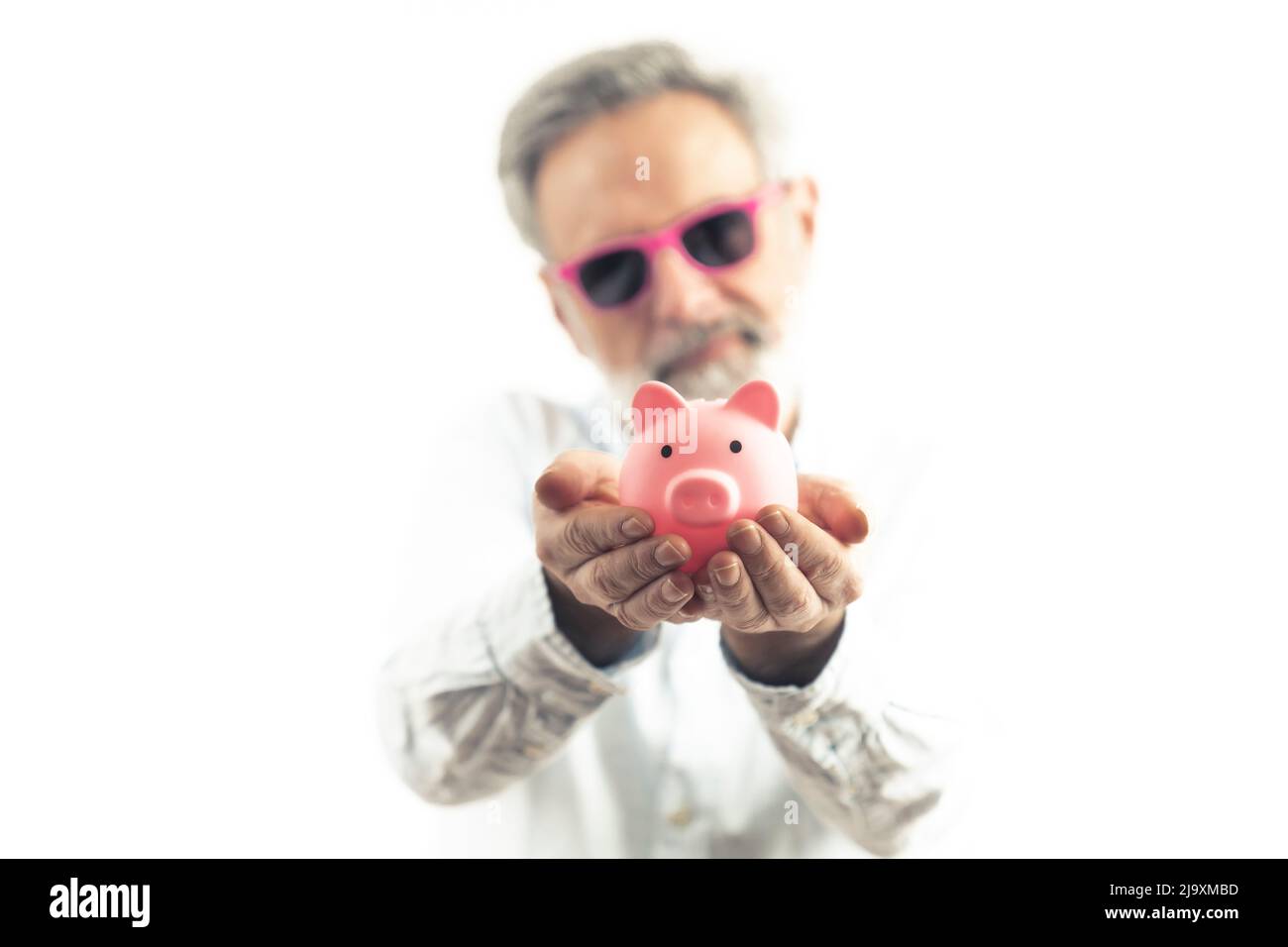 Flippiger kaukasischer älterer Mann mit grauem Haar in rosa Sonnenbrille, der das Sparschwein hält und es der Kamera zeigt. Sparkonzept und Happy Retirement. Hochwertige Fotos Stockfoto