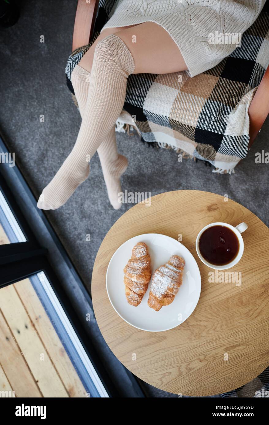 Draufsicht auf die Beine einer jungen Frau in der Nähe des Fensters im modernen Scheunenhaus. Mit Croissants und einer Tasse Tee auf den Tisch legen. Stockfoto
