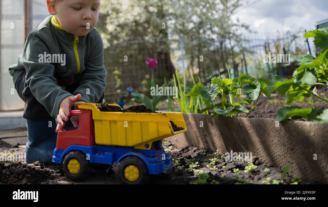 Aktiver Junge Vorschulkinder mit blonden Haaren, die mit einem Plastikwagen im Garten spielen. Das Kind lernt ohne Aufsicht eines Erwachsenen spielen. Glücklich und vorsichtig Stockfoto