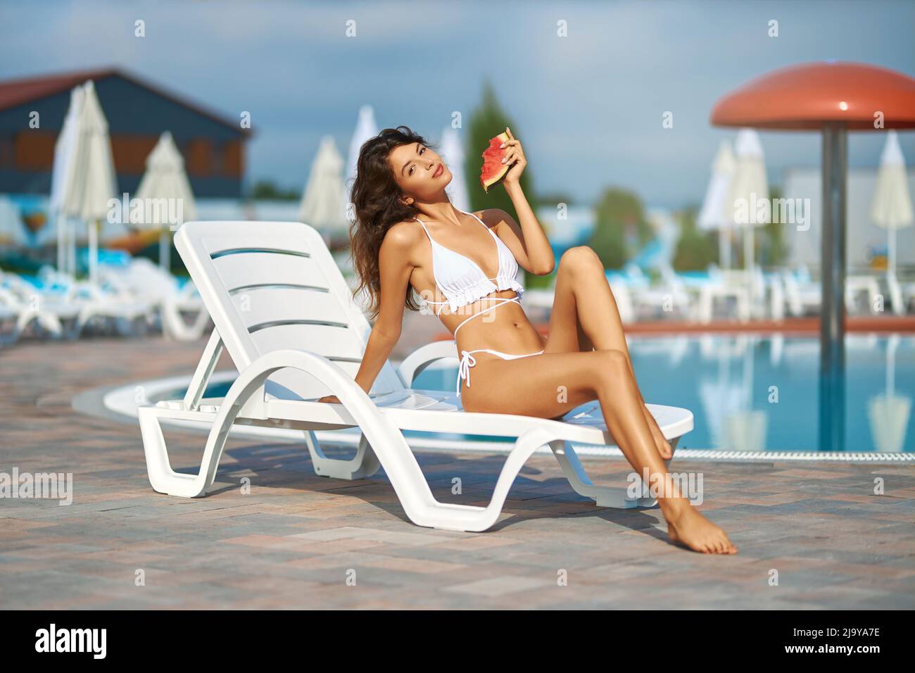 Hübsche weibliche Touristen posieren, während Sonnenbaden auf dem Liegesessel draußen. Vorderansicht eines anmutigen Mädchens, das die Kamera anlächelt, während es Wassermelone isst, mit verschwommenem Pool im Hintergrund. Konzept der Ruhe. Stockfoto