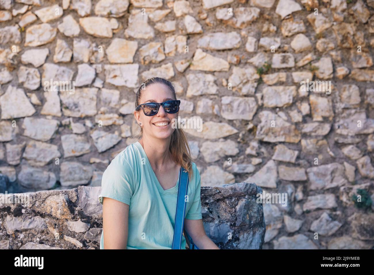 Hübsches blondes Mädchen lächelt mit Sonnenbrille und Steinwand Hintergrund Stockfoto