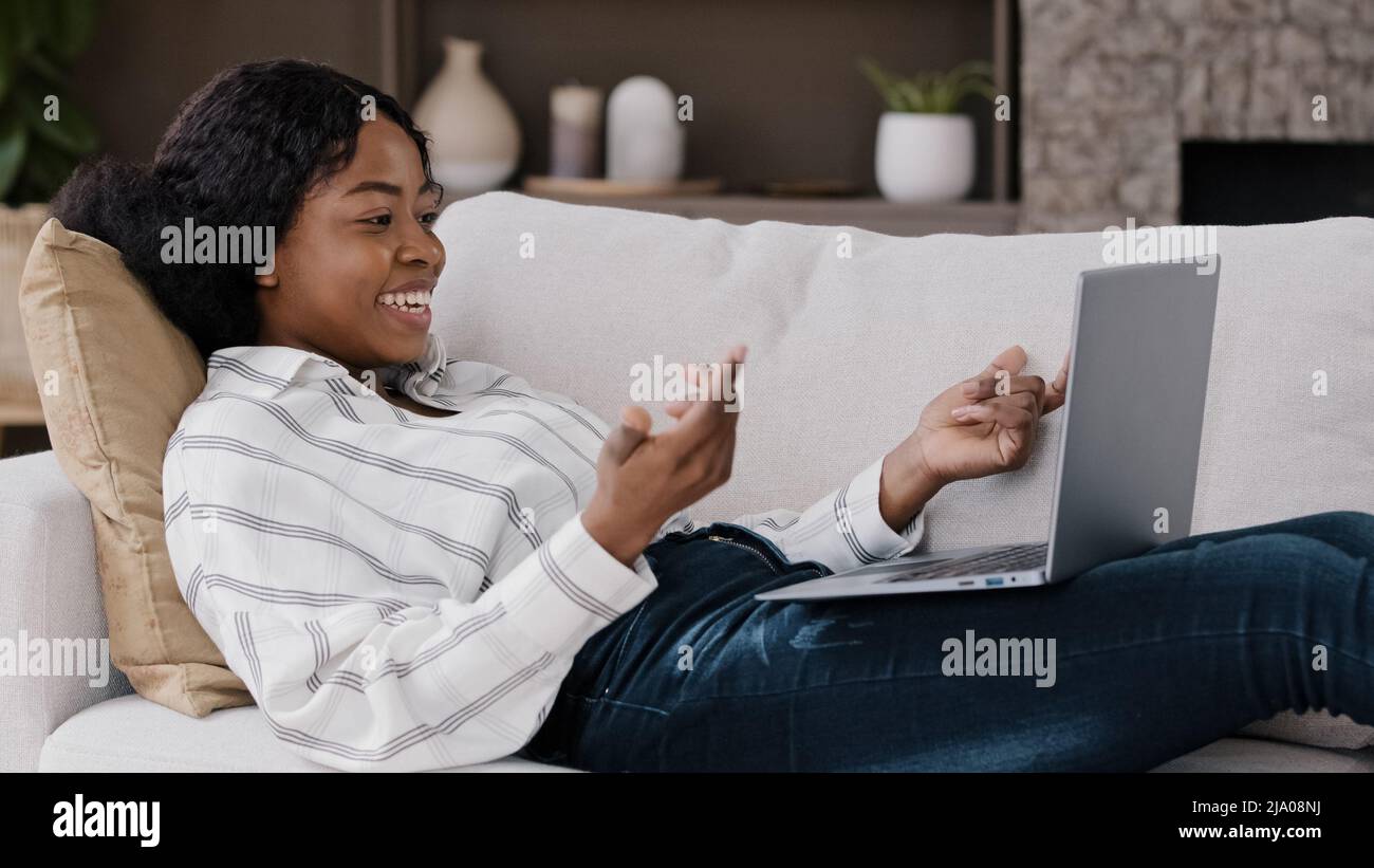 Afrikanische Frau, die zu Hause auf dem Sofa liegt, schaut auf den Laptop-Bildschirm, grüßt Hallo, macht einen Videoanruf und unterhält sich mit der Familie eines Freundes aus der Ferne Stockfoto