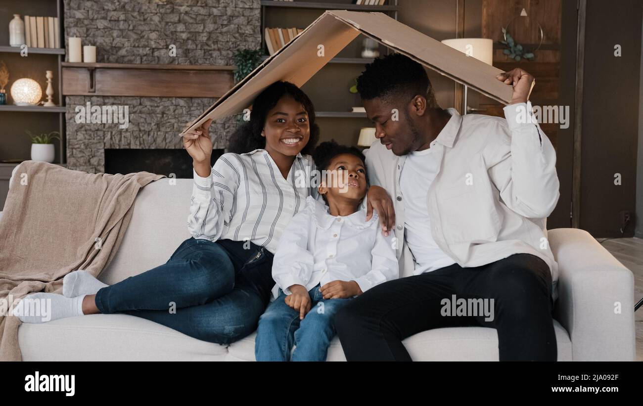 afroamerikanische Familieneltern mit Tochter kleines Mädchen adoptierte Kind sitzen auf dem Sofa unter Kartondach träumen von eigenen Haus Umzug nach Hause Stockfoto