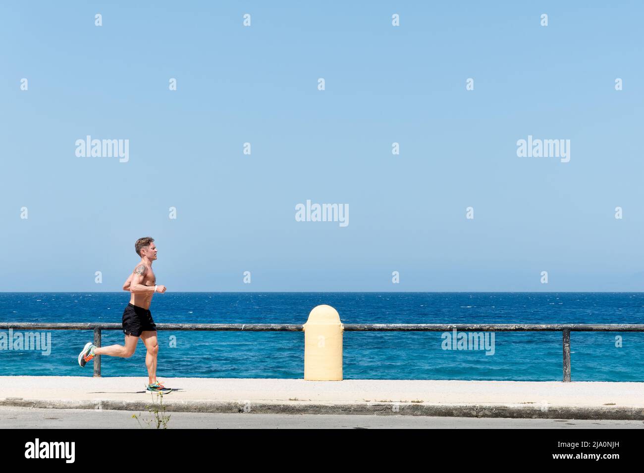 Ein Jogger oder Läufer, der hart an einer Straße am Meer trainiert und bei heißem Sonnenschein bis zur Taille entkleidet ist. Im Hintergrund ist ein türkisfarbenes Meer zu sehen Stockfoto