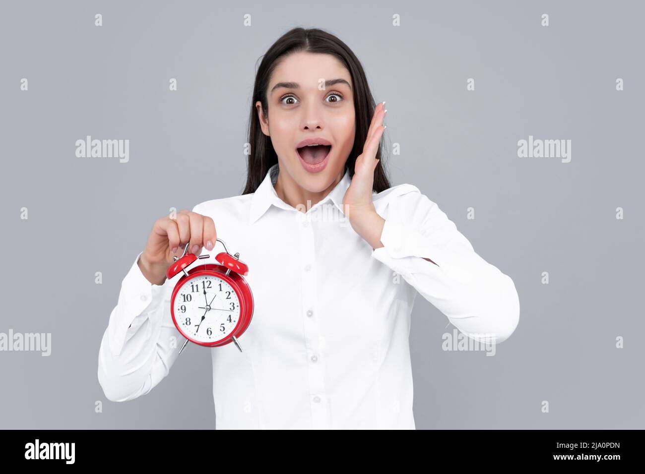 Frau hält eine Uhr. Emotionales junges weibliches Gesicht. Porträt einer jungen überraschten Frau mit Wecker auf grauem Hintergrund. Stockfoto