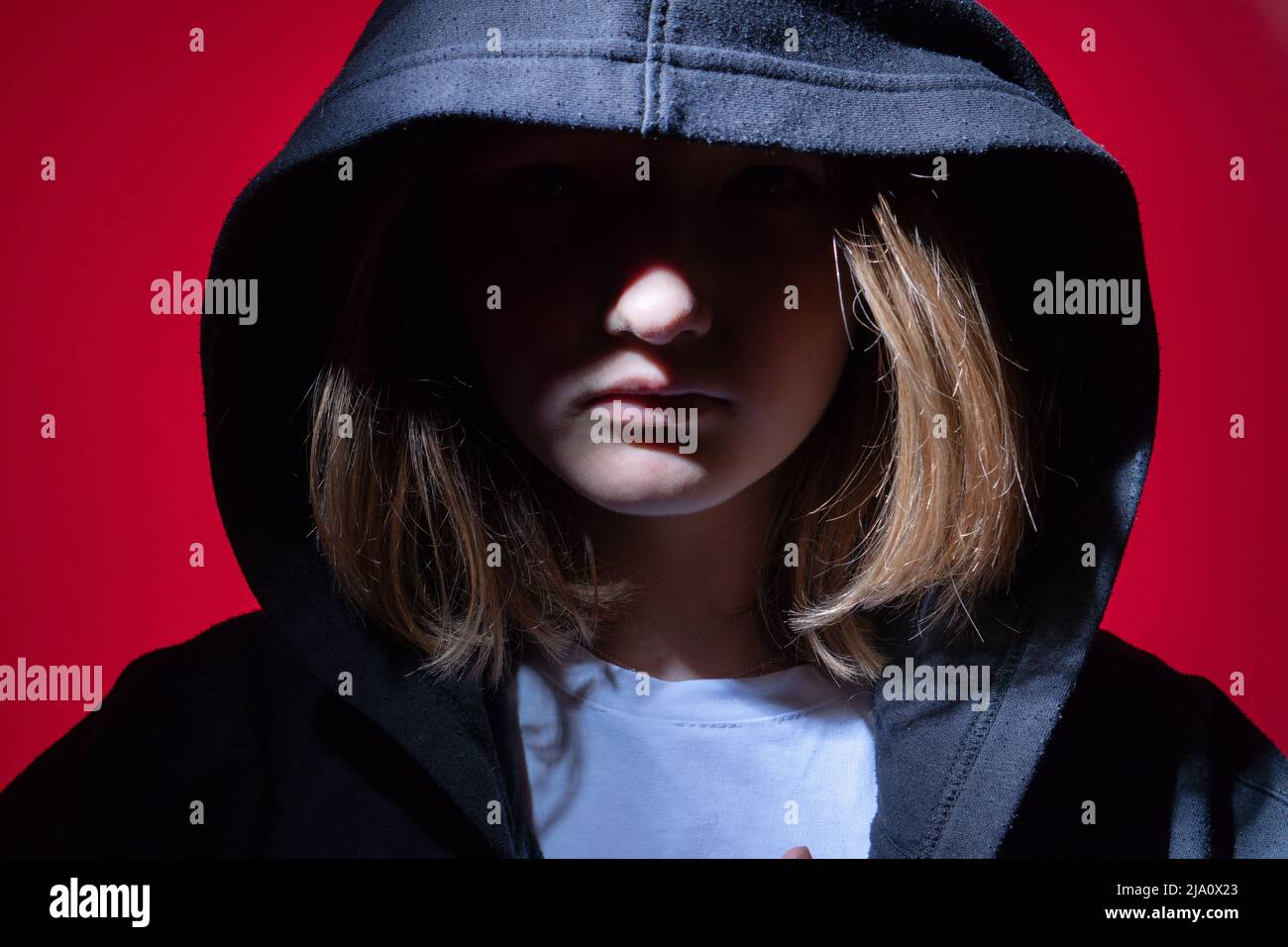 Ein Mädchen im Teenageralter in einem schwarzen Hoodie mit einer Kapuze auf rotem Hintergrund. Das Gesicht ist geschlossen. Das Konzept der Probleme bei Jugendlichen und die Schwierigkeiten der t Stockfoto