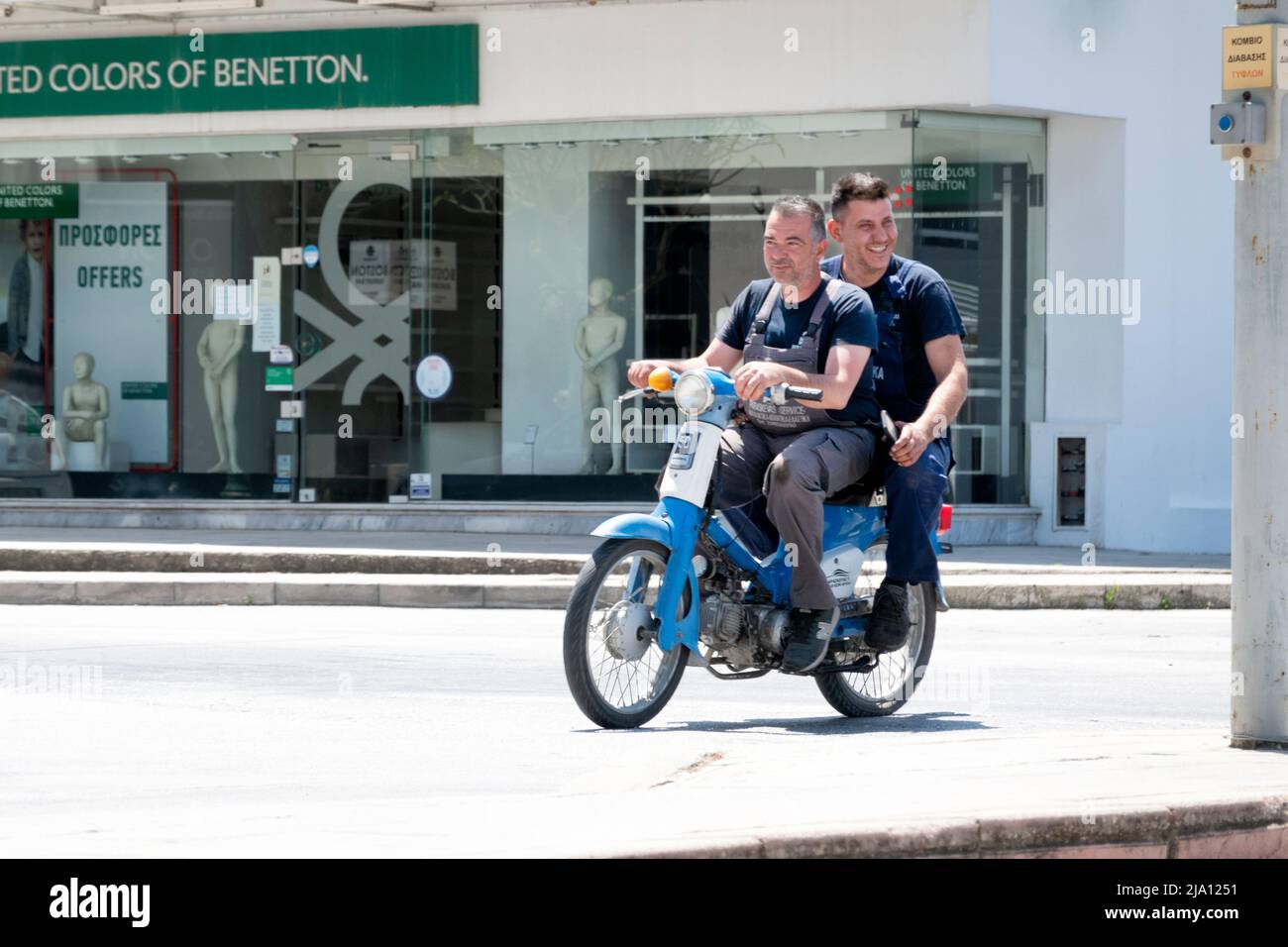 Zwei Arbeiter, die auf einem kleinen Motorrad auf einer befahrenen Straße in Rhodos, Griechenland, unterwegs sind. Die Männer sind gut gelaunt und immer noch in ihrer Arbeit Overalls Stockfoto