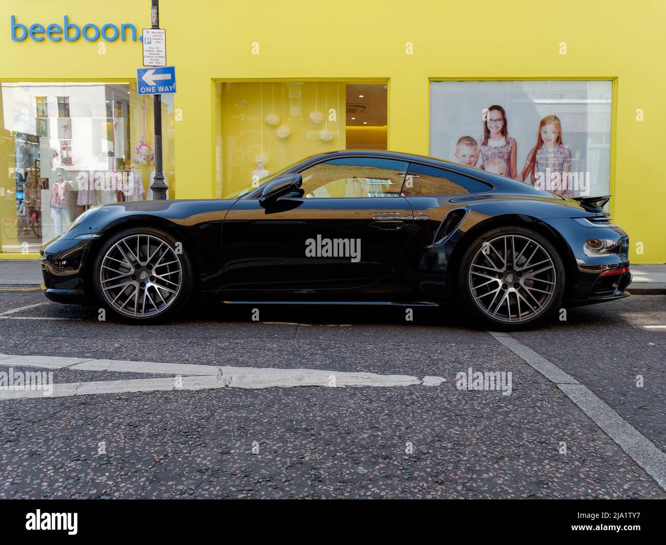 London, Greater London, England, Mai 14 2022: Schwarzer Porsche-Sportwagen vor gelbem Ladenhintergrund mit Straßenmarkierungen im Vordergrund. Stockfoto