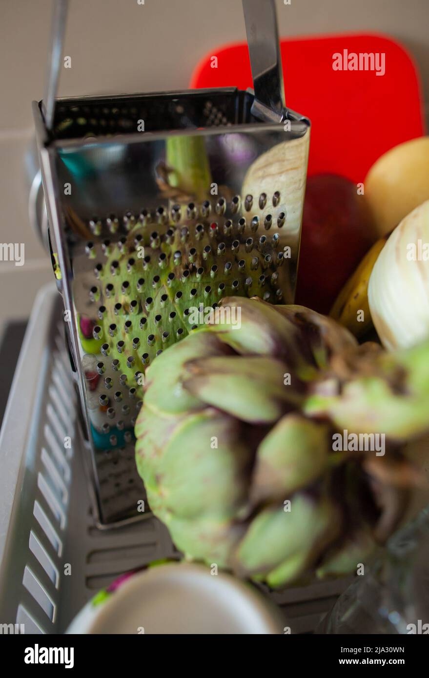 Gereinigtes Gemüse und Geschirr auf grauem Kunststoffschüssel-Trockengestell Closeup Artichoke Reflexion in silberner Reibe auf dem Geschirrtrocknungsgestell mit anderem Gemüse Stockfoto