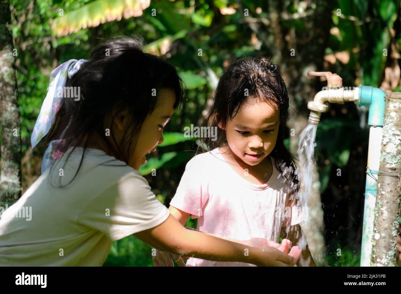 Zwei kleine Schwestern waschen sich die Hände am Wasserhahn im Freien. Zwei niedliche Mädchen drehen den Wasserhahn zu, um sich im Park die Hände zu waschen. Stockfoto