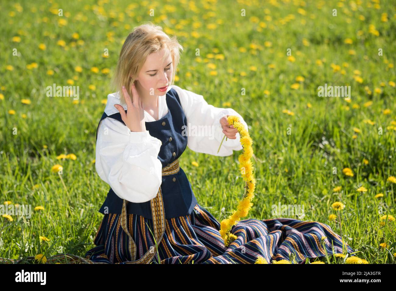 Junge Frau in Nationalkleidung mit gelbem Dandelion-Kranz im Frühlingsfeld. Frühling Stockfoto