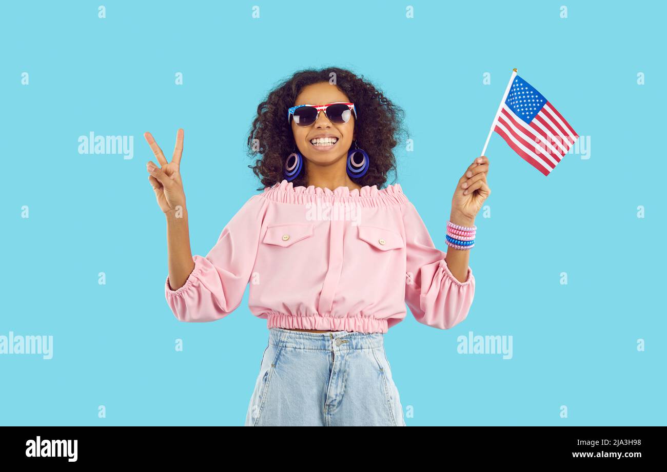 Glückliches dunkelhäutig jugendliches Mädchen, das auf hellblauem Hintergrund das Siegeszeichen mit der amerikanischen Flagge zeigt. Stockfoto