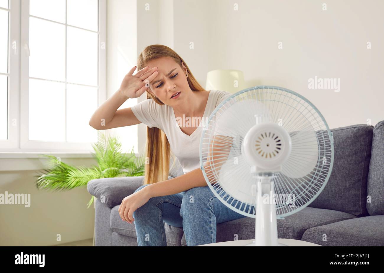 Erschöpfte überhitzte Frau wird durch kalte Luft gekühlt, die auf dem Sofa vor dem elektrischen Ventilator sitzt. Stockfoto