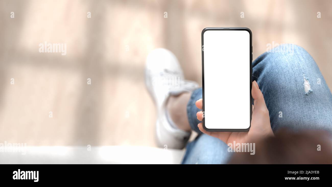 Mockup-Bild von oben mit einer Frau, die ein Mobiltelefon mit leerem weißen Bildschirm hält. Frau in Jeans sitzt mit gekreuzten Beinen und hält das Handy in den Händen mit b Stockfoto
