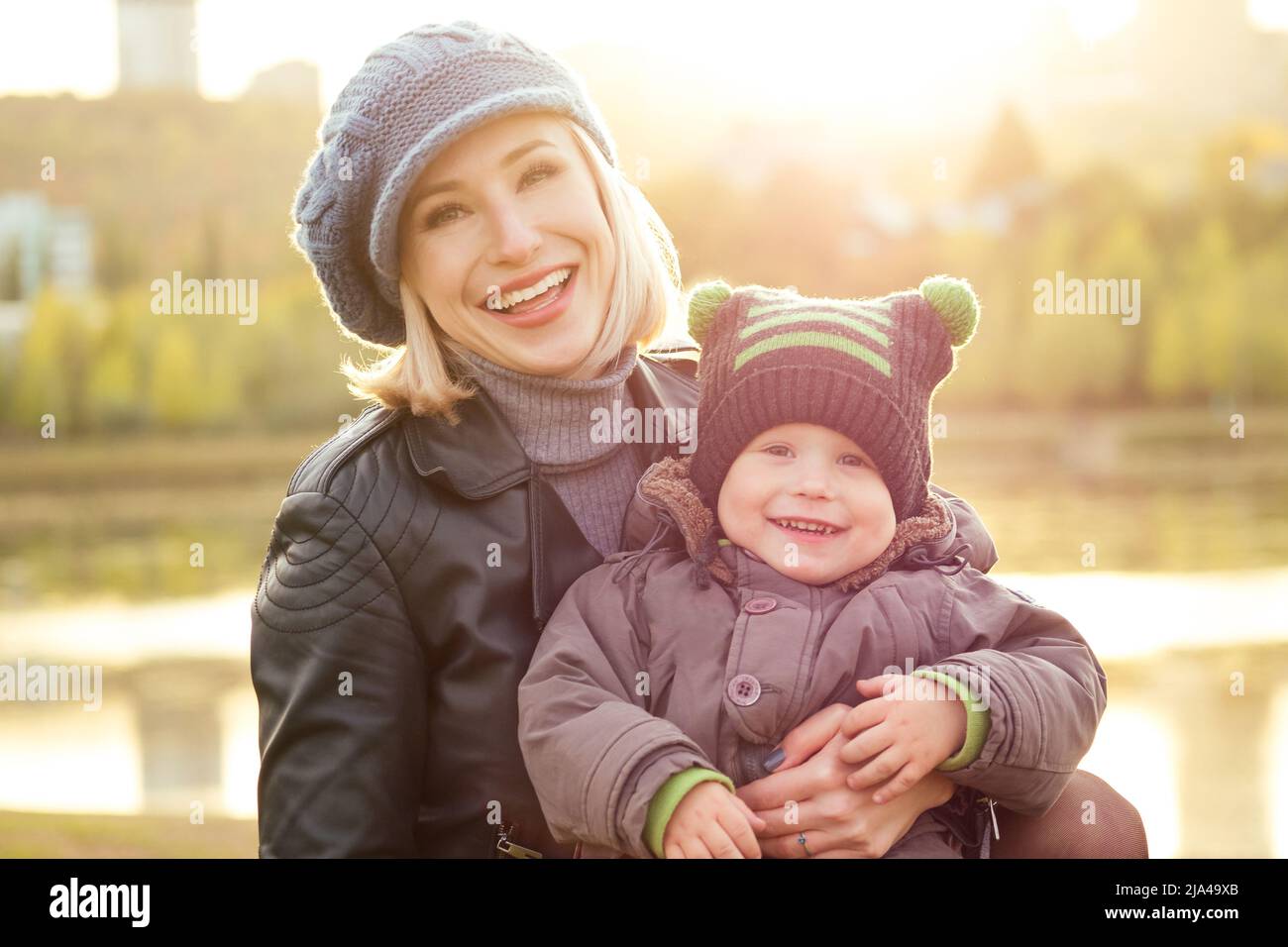 Mutter in einer warmen Jacke und Strickmütze umarmt ihren kleinen Jungen zu Fuß im Herbst Park. Stilvolle blondwoman mit einem Kind vor der Kulisse der Stockfoto