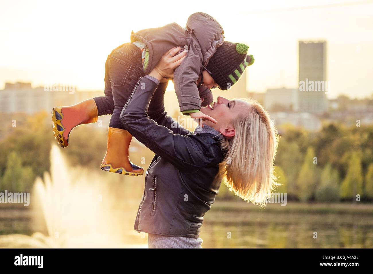 Mutter in einer warmen Jacke und gestrickten Hut werfen ihren kleinen Jungen zu Fuß im Herbst Park. Stilvolle blondwoman mit einem Kind vor der Kulisse der Stockfoto