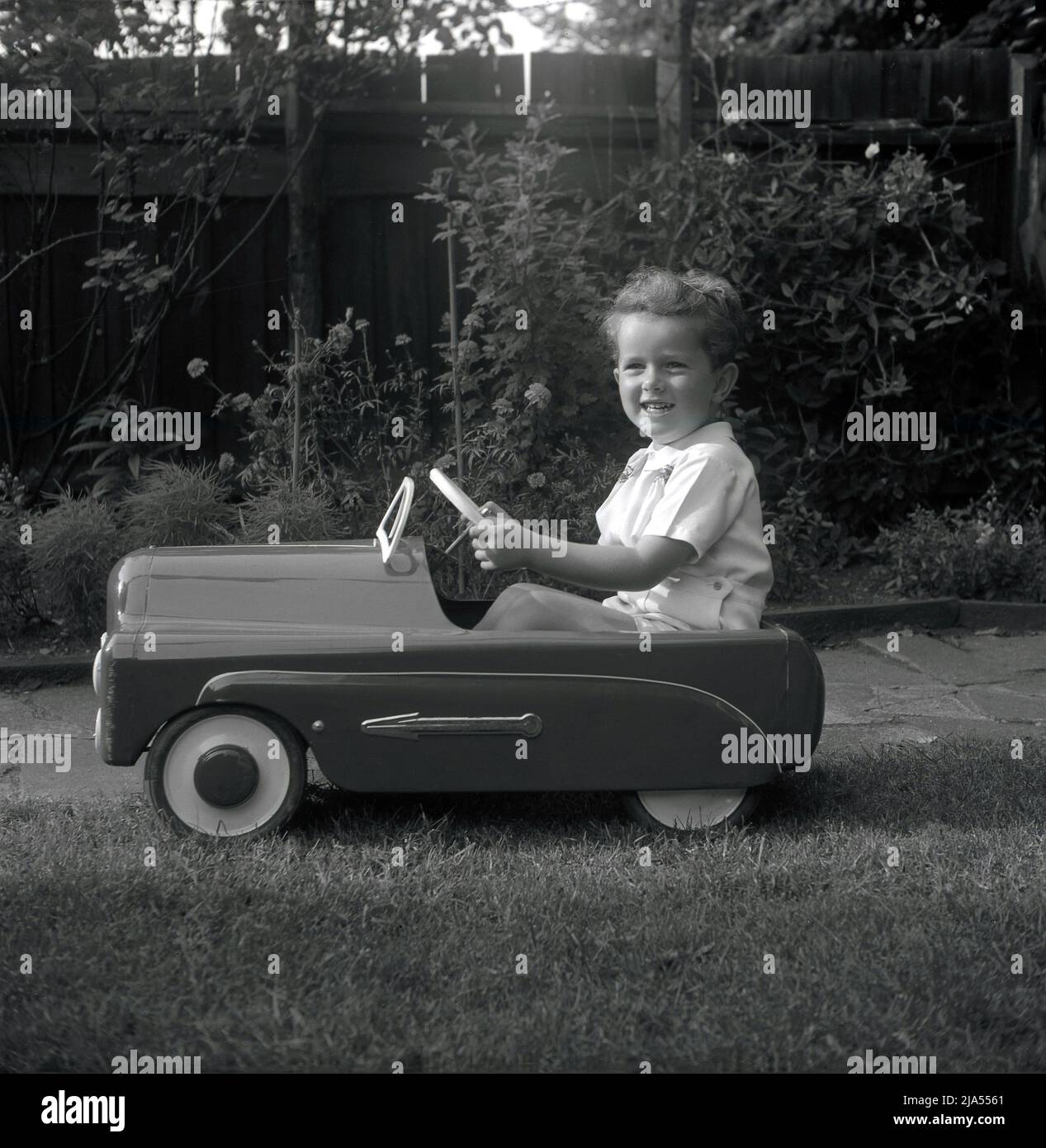 1953, historisch, draußen in einem Garten, ein kleiner Junge, der in seinem fahrbaren Spielzeugpedal der damaligen Zeit sitzt, ein britischer Tri-Ang 'Thirty B'. Aus gepresstem Stahl gefertigt, hatte das Spielzeugauto eine Hochglanz-Emaille-Oberfläche mit Chrompfeilen an der Seite. Tri-Ang war der Markenname von Line Bros. Ltd, einem britischen Unternehmen, das in dieser Zeit das größte Spielzeug- und Babywagenhersteller der Welt war. Stockfoto