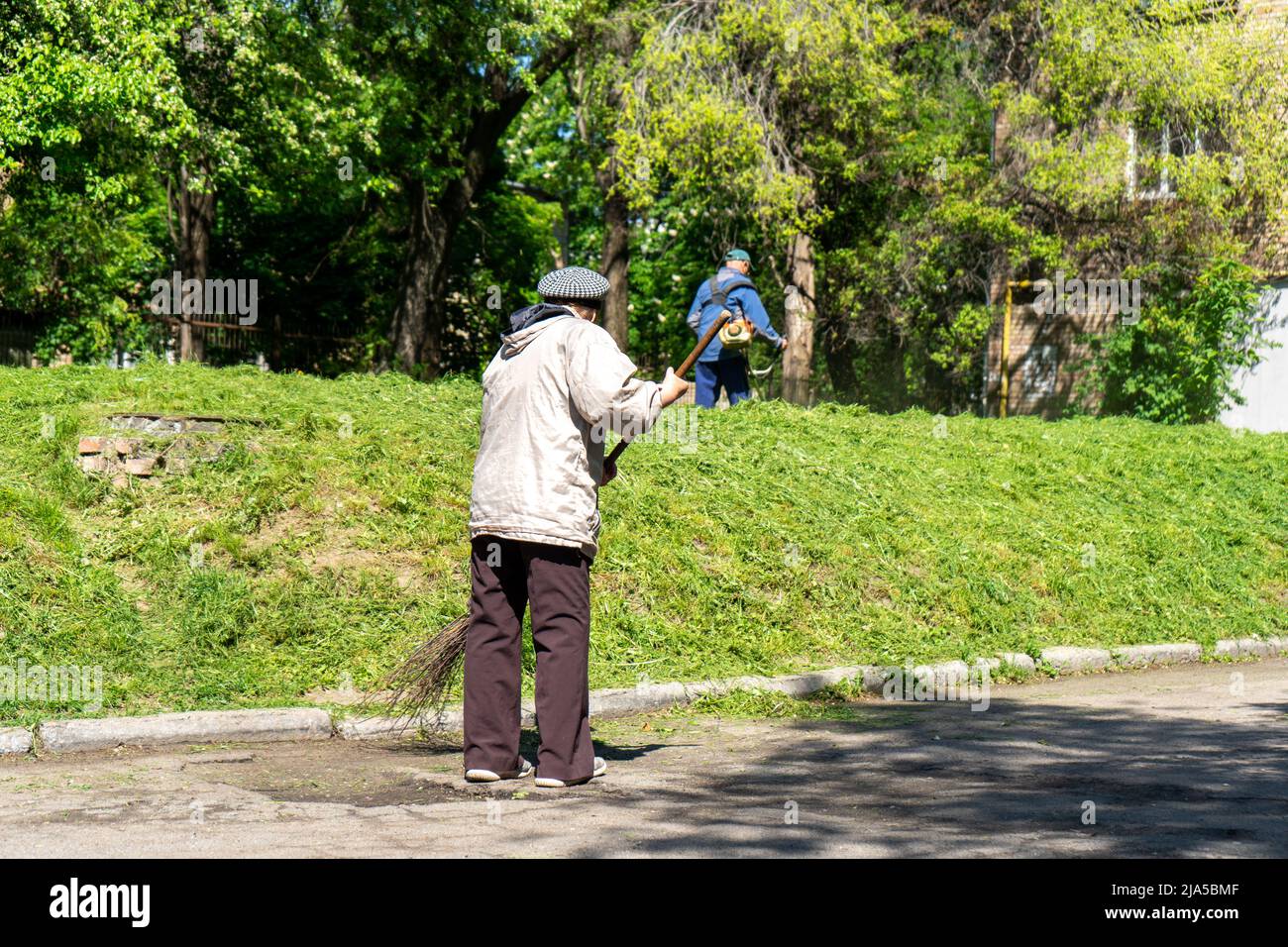 Alte Frau, die die Straße in der Stadt fegen. Mann im Hintergrund mäht das Gras Stockfoto
