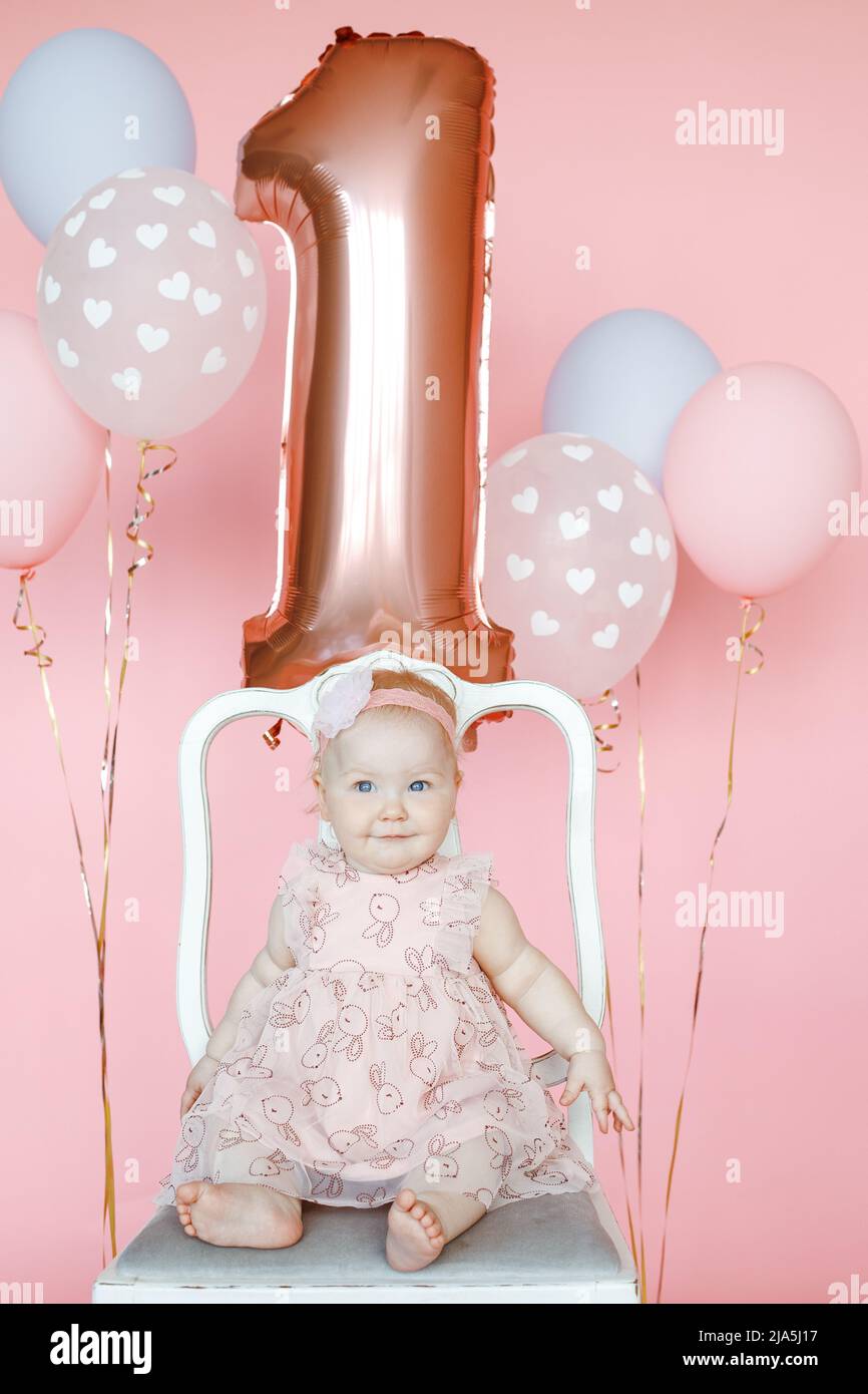 Portrait von schönen ein Jahr alten Baby Mädchen im Urlaub Kleid sitzt auf einem Stuhl unter Helium Ballons auf rosa Hintergrund. Erste Geburtstagsfeier, rosa Stockfoto