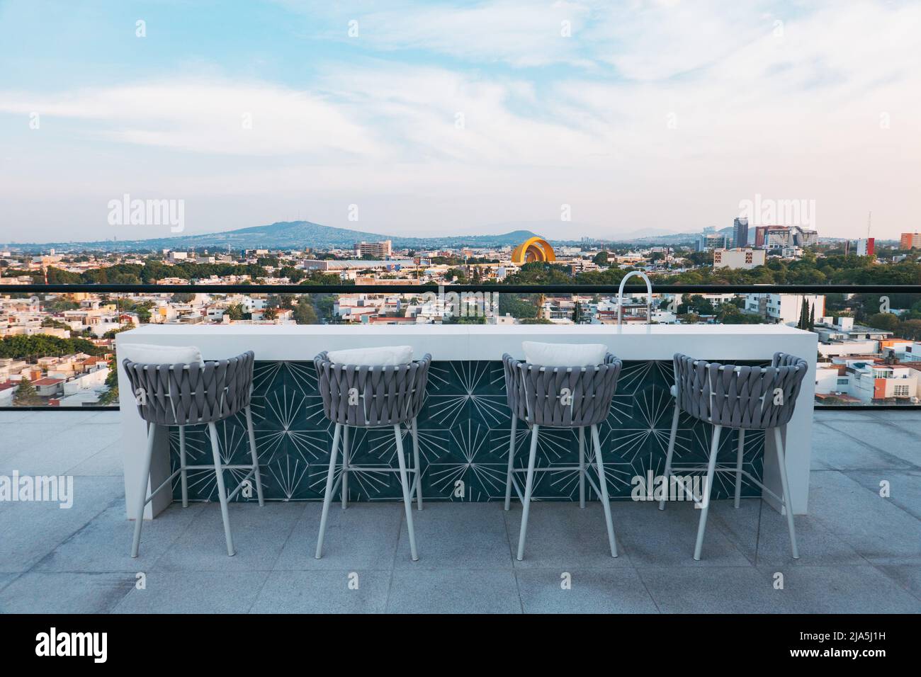 Hocker und ein Bartisch blicken vom Dach eines neuen Apartmentkomplexes in Jardines del Bosque, Guadalajara, Mexiko, auf die Skyline Stockfoto