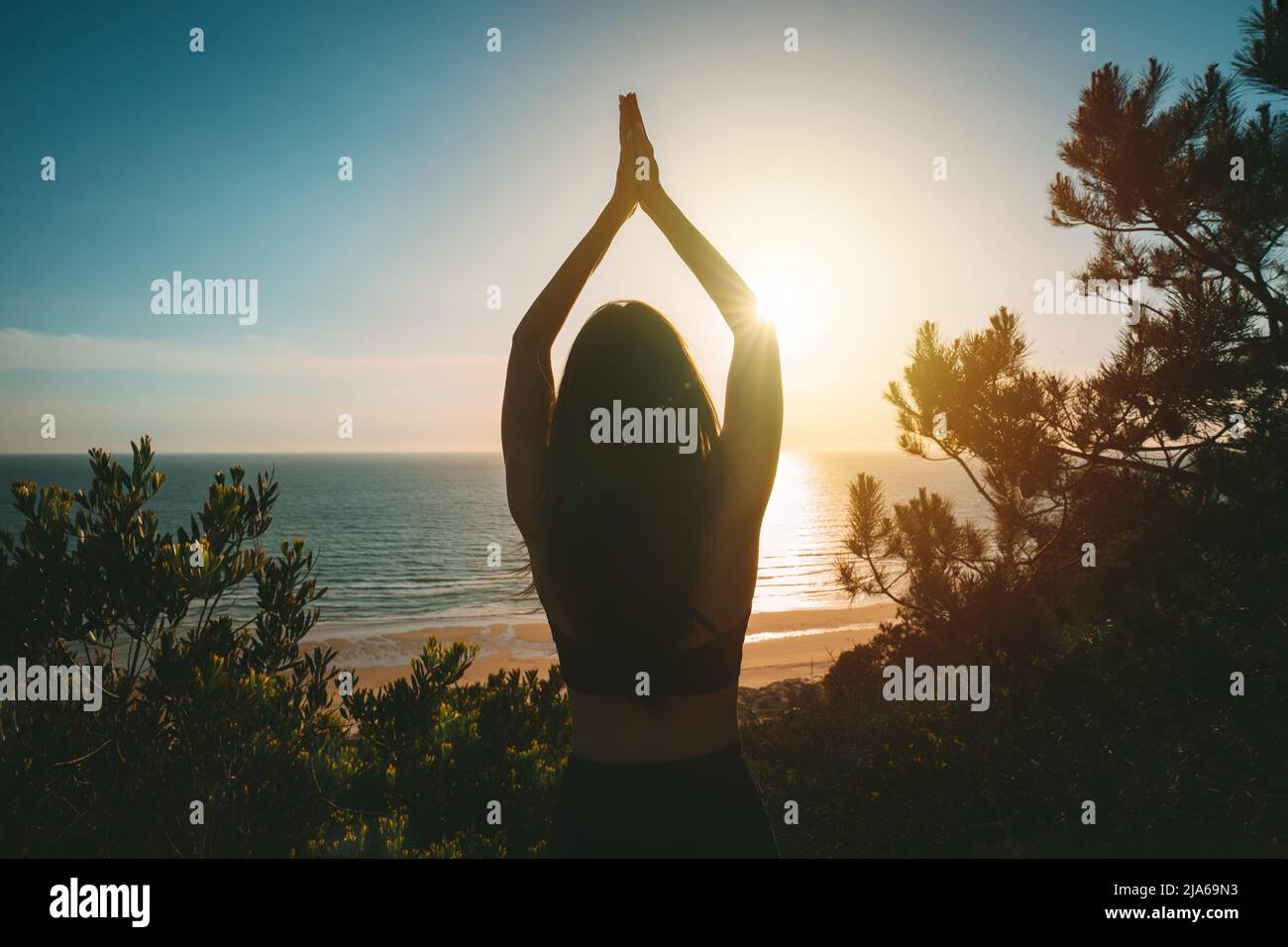Silhouette einer jungen, gesunden Frau, die Yoga praktiziert, mit Blick auf den Ozean bei Sonnenuntergang. Rückansicht einer Frau, die mit den Händen in Baumhaltung über dem Kopf steht Stockfoto