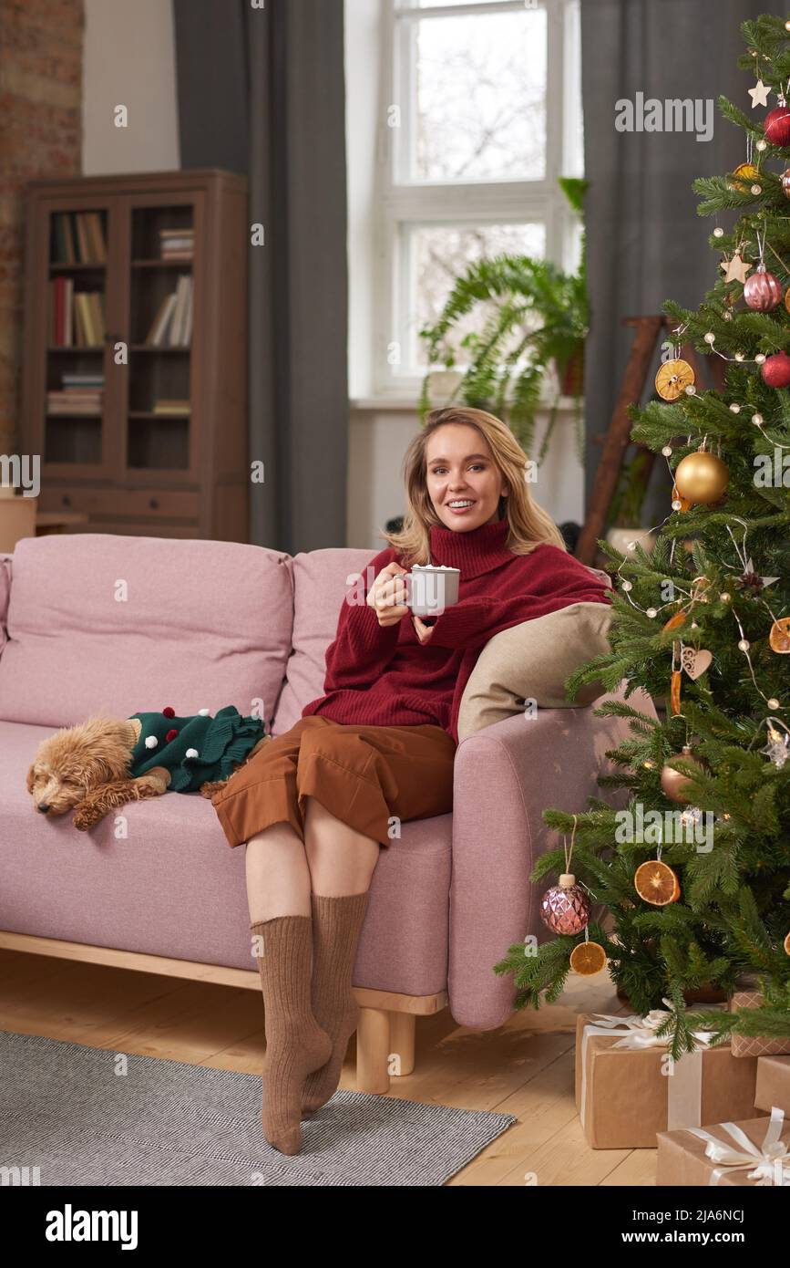 Vertikale Vollaufnahme der stilvollen jungen Frau in warmen Kleidern verbringen Weihnachtsmorgen zu Hause trinken heiße Schokolade Milch im Wohnzimmer Stockfoto