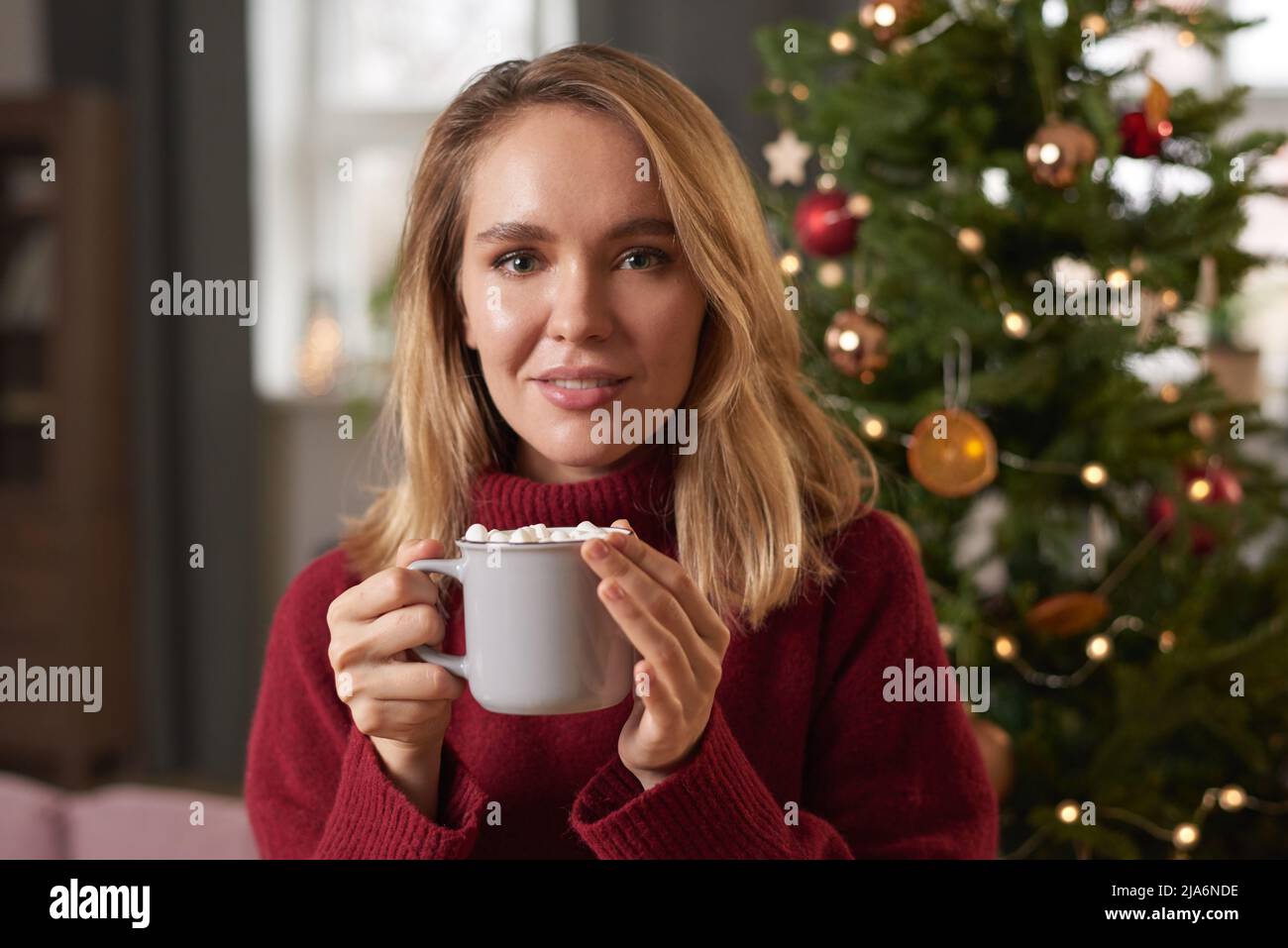 Mittellange Nahaufnahme einer attraktiven jungen Frau mit blondem Haar in einem warmen Pullover, die heißen Kaffee mit Marschmalow trinkt Stockfoto