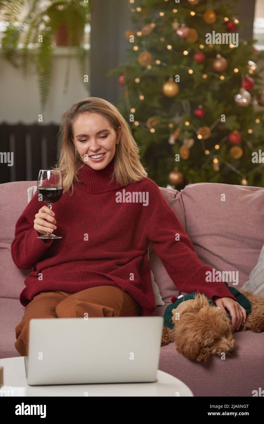 Porträt einer fröhlichen jungen Frau, die auf dem Sofa im Wohnzimmer sitzt, das zu Weihnachten dekoriert ist, Rotwein trinkt, Hund streichelnd und mit Freunden plaudert Stockfoto