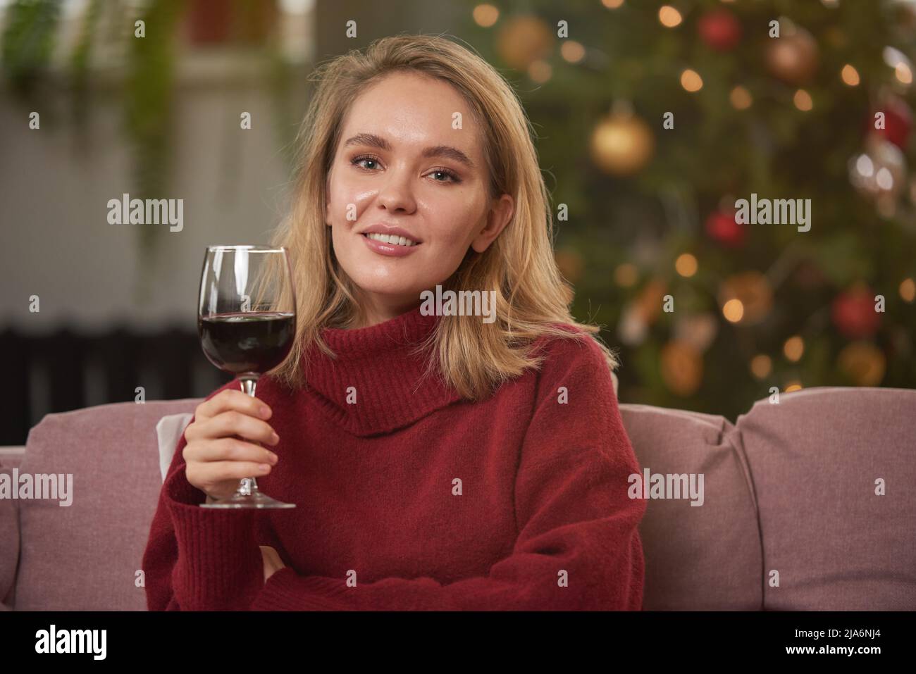 Mittelgroßes Nahaufnahme-Porträt einer attraktiven Frau mit warmem Pullover, die auf dem Sofa im zu Weihnachten dekorierten Wohnzimmer sitzt und ein Glas Rotwein hält Stockfoto